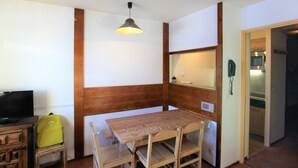 A compact room with a wooden table and chairs, a television on a wooden cabinet, a hanging light fixture, and a telephone on the wall.
