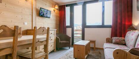 Una habitación acogedora con paredes de madera, un televisor montado en la pared, un sofá, una silla, una mesita y una ventana con cortinas.