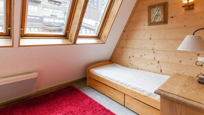Une petite pièce aux murs inclinés, avec un lit simple, un tapis rouge et un mur en bois orné d’un tableau et d’une lampe.