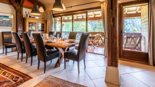 A dining area with a wooden table and chairs, large windows, and a view of the outdoors.