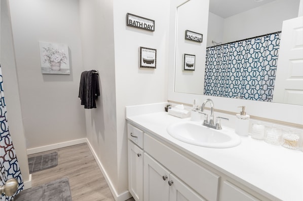 A bathroom with a white sink, mirror, and towel rack. There are framed signs on the wall and a shower curtain with a geometric pattern. - Edmond Oasis: Luxury Retreat w/ Massage Chair and Infinity Game Table (Edmond)