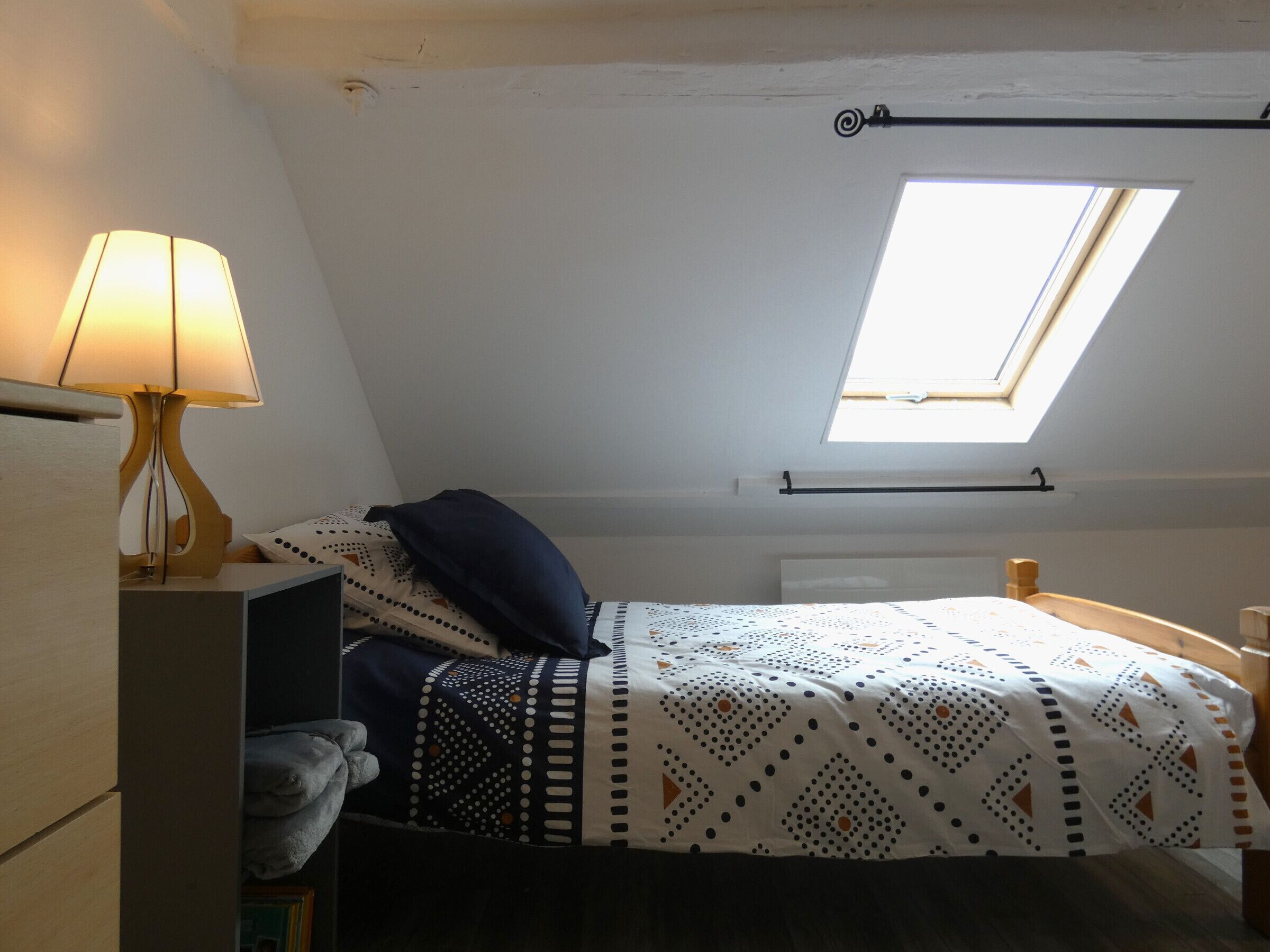 A small, cozy bedroom with a bed, a nightstand, a lamp, and a skylight.