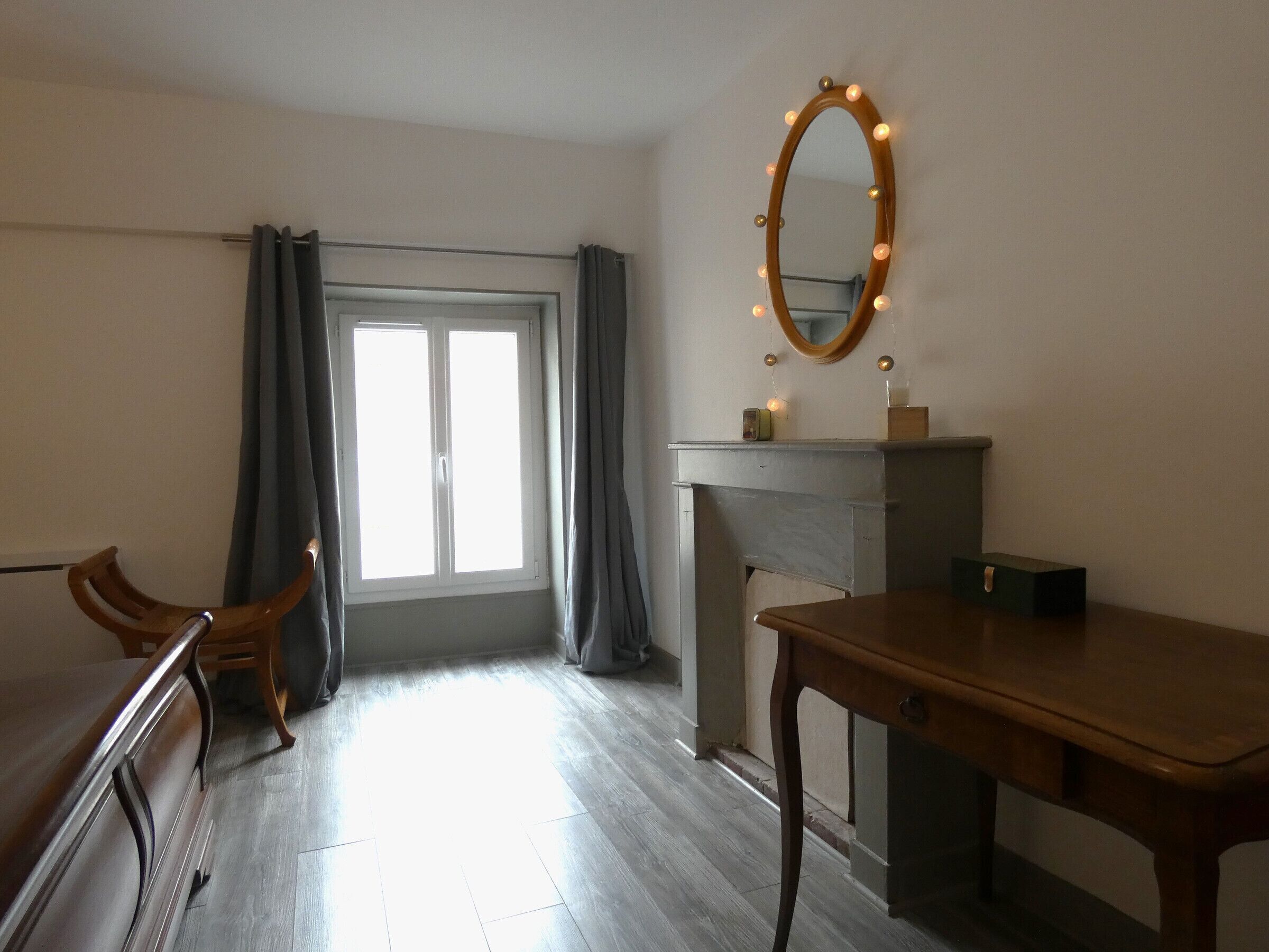 Une pièce avec un bureau en bois, une chaise, un miroir avec des lumières, une cheminée et une fenêtre avec des rideaux.
