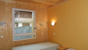 A room with two beds, wooden walls, a window with a view of a balcony, and a wall-mounted light fixture. - Charming 2-Bed Apartment, Wi-Fi, Near Ski Lifts & Center, Les Gets (Les Gets)