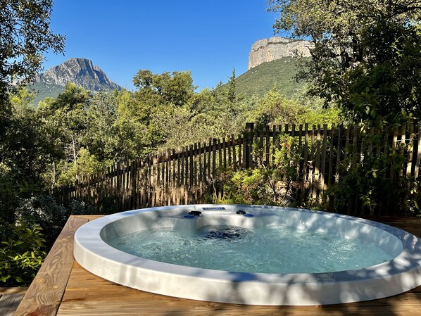 Un jacuzzi en un jardín con terraza de madera, rodeado de un cerco de madera y exuberante vegetación, con montañas al fondo.