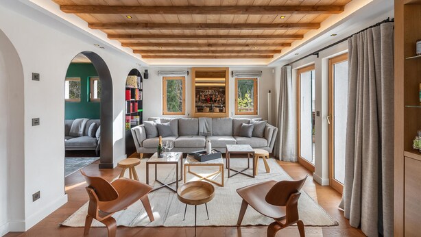 Ein modernes Wohnzimmer mit Holzboden, einem Ecksofa und einem Couchtisch. Ein Bücherregal und ein Fenster mit Blick auf Bäume sind ebenfalls vorhanden.