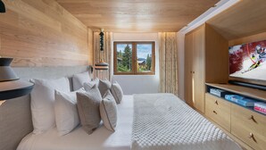 Ein Schlafzimmer mit Holzboden, einem Bett mit weißer Bettwäsche, einem Fernseher rechts und einem Fenster mit Blick auf Bäume und Häuser.