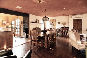 A spacious dining area with a wooden table and chairs, a hanging light fixture, and a view into a kitchen area.