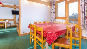 Uma sala de jantar com uma mesa posta para seis, cadeiras de madeira, toalha vermelha e vista pela janela com cortinas.