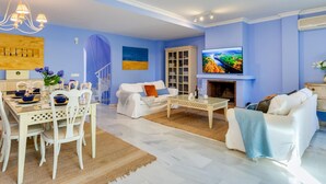 A living room with a white sofa, a coffee table, and a TV. There is a fireplace and a dining area with white chairs and a table. - Luxury Beachfront Villa with Private Terrace & Pools in Puerto Banús (Marbella)