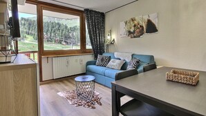 A modern living room with a blue sofa, a dining table, and a view of a mountainous landscape through a large window. - Renovated studio for 4 pers at the foot of the slopes (La Plagne)