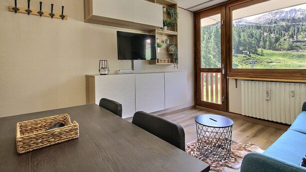 A dining area with a wooden table, chairs, a TV mounted on a cabinet, and a view of a mountainous landscape through a large window. - Renovated studio for 4 pers at the foot of the slopes (La Plagne)