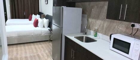 A compact hotel room with a bed, a kitchenette, a microwave, and a ceiling fan.