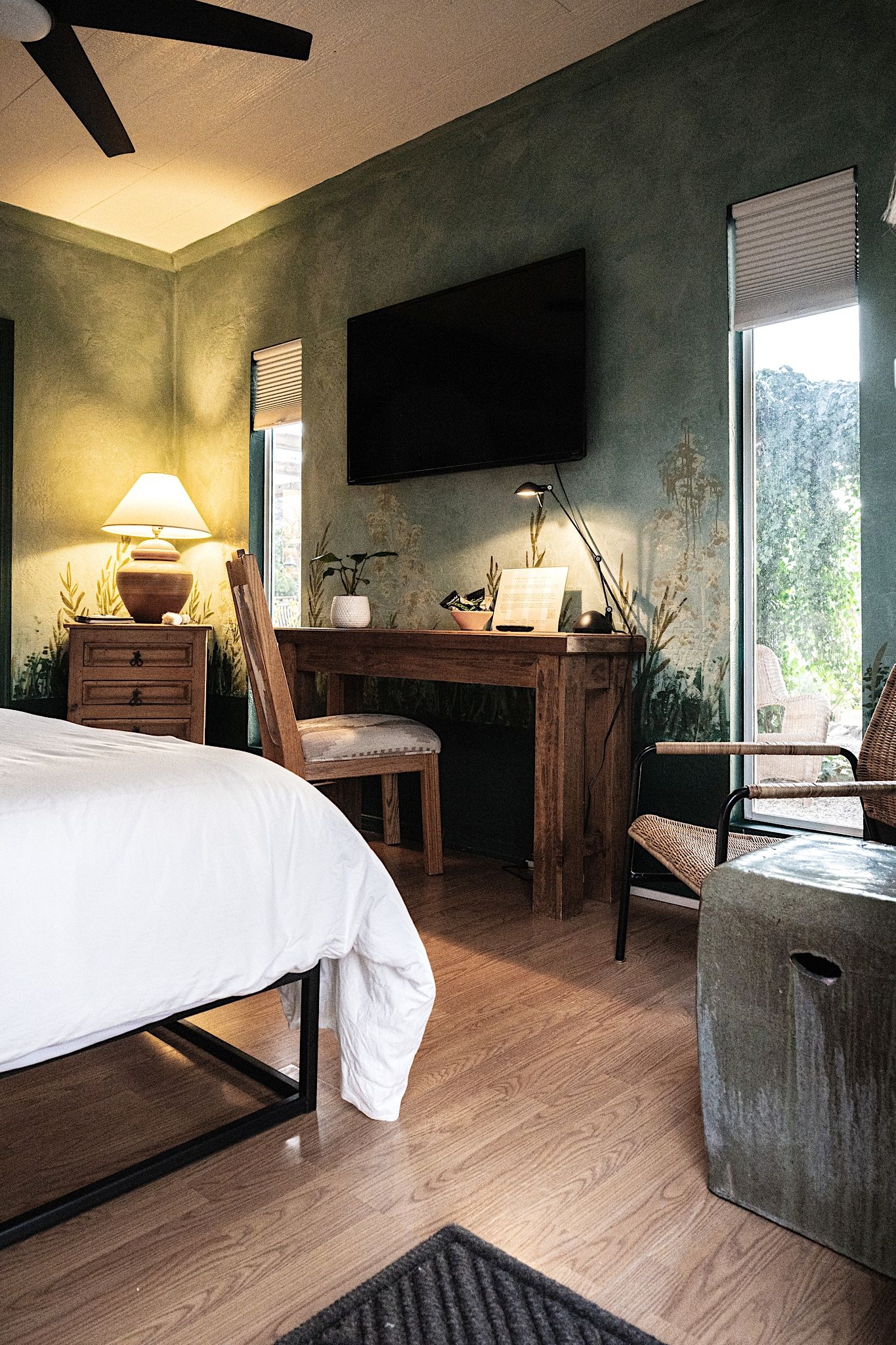 A bedroom with a bed, desk, and chairs, a television on the wall, and a window with a view.