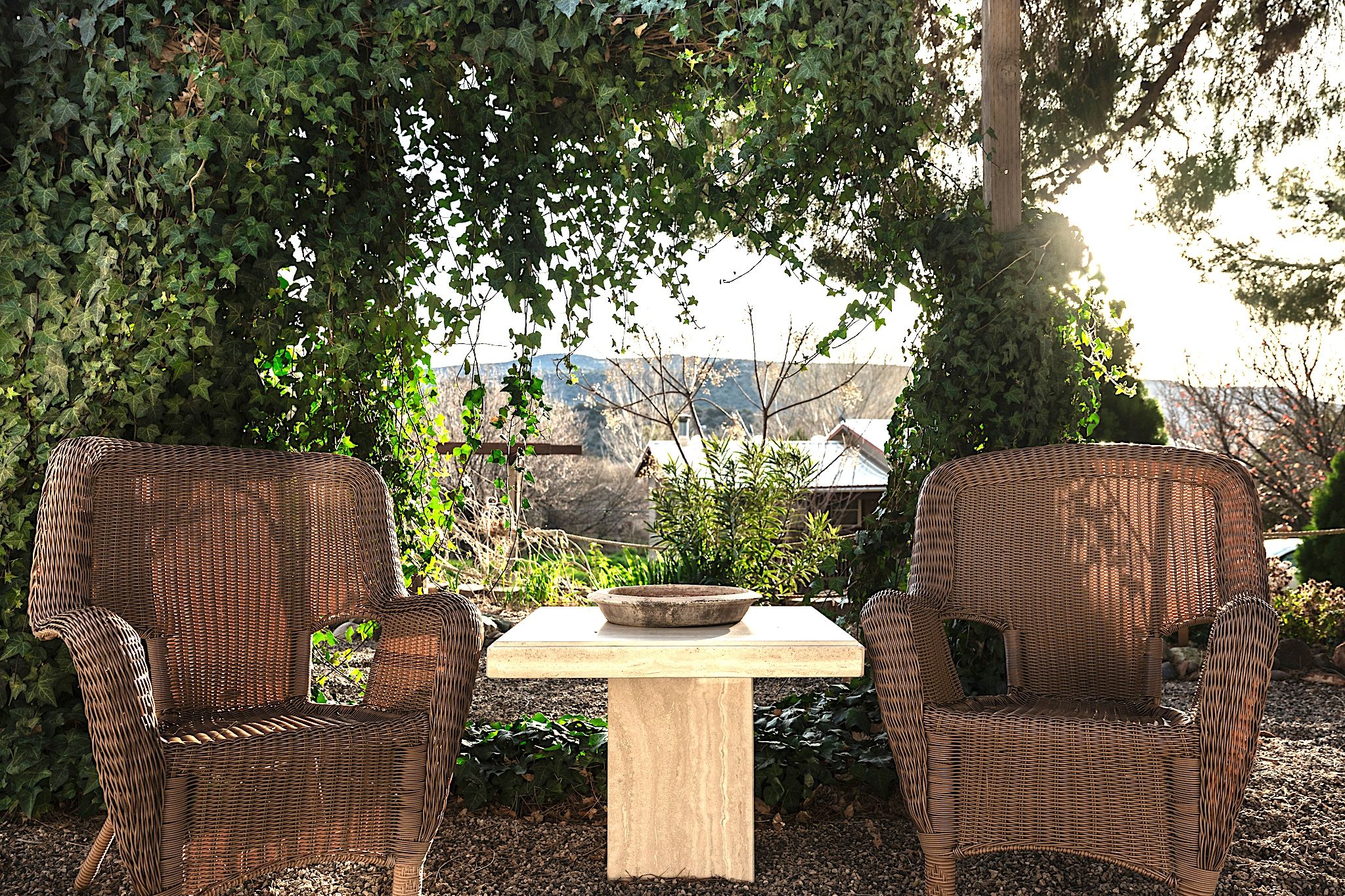 Deux chaises en osier et une table avec un saladier, disposés à l’extérieur, avec vue sur une maison et des arbres.
