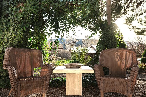 Dos sillas de mimbre y una mesa con un cuenco, ubicados al aire libre con vista a una casa y árboles.