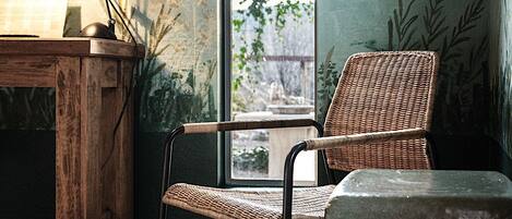 A room with a wooden desk, a wicker chair, a large window, and a view of greenery outside.