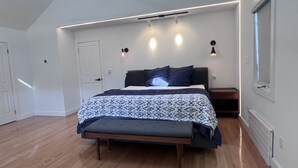 A modern bedroom with a large bed, a bench at the foot, a nightstand, and a wall-mounted light fixture. - Family Friendly Villa with Pool (Queens)