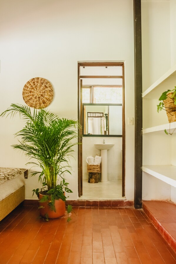 A narrow hallway with a potted plant, a wicker wall hanging, and a view into a bathroom with a sink and shelves.