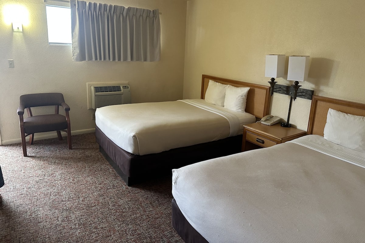 A hotel room with two beds, a chair, an air conditioning unit, a lamp, and a window with curtains.