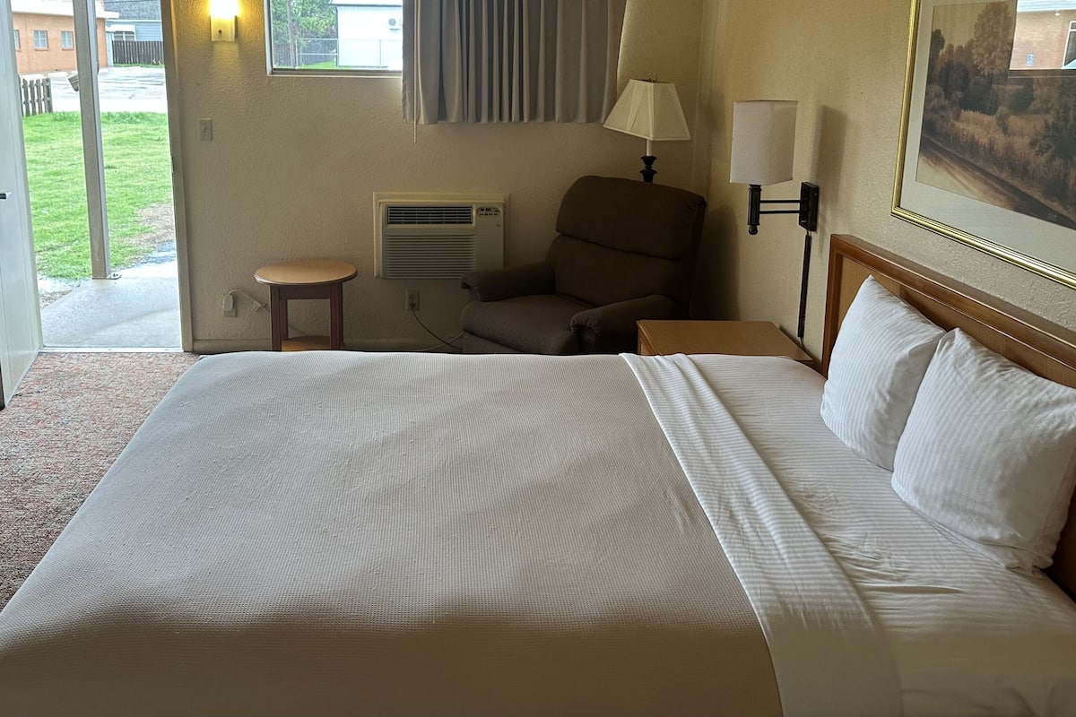 A hotel room with a large bed, a chair, a table, a lamp, and a painting on the wall.
