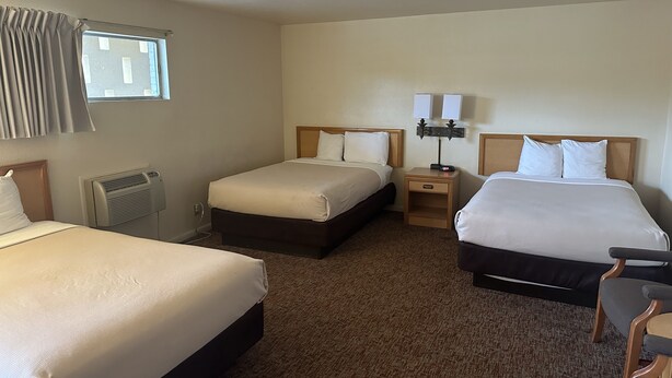 A hotel room with two single beds, a nightstand with a lamp, a heater, and a window with curtains. - Big Bend Traveler's Inn (Great Bend)