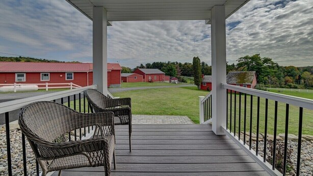 A covered outdoor area with wicker chairs, a view of red buildings, and a grassy landscape.