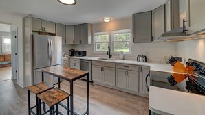 A modern kitchen with light grey cabinets, a stainless steel refrigerator, a wooden table, and a large window providing natural light.