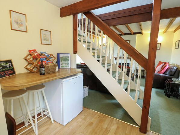 A kitchen area with a wooden counter, white stools, a small refrigerator, and a staircase leading to a living room.