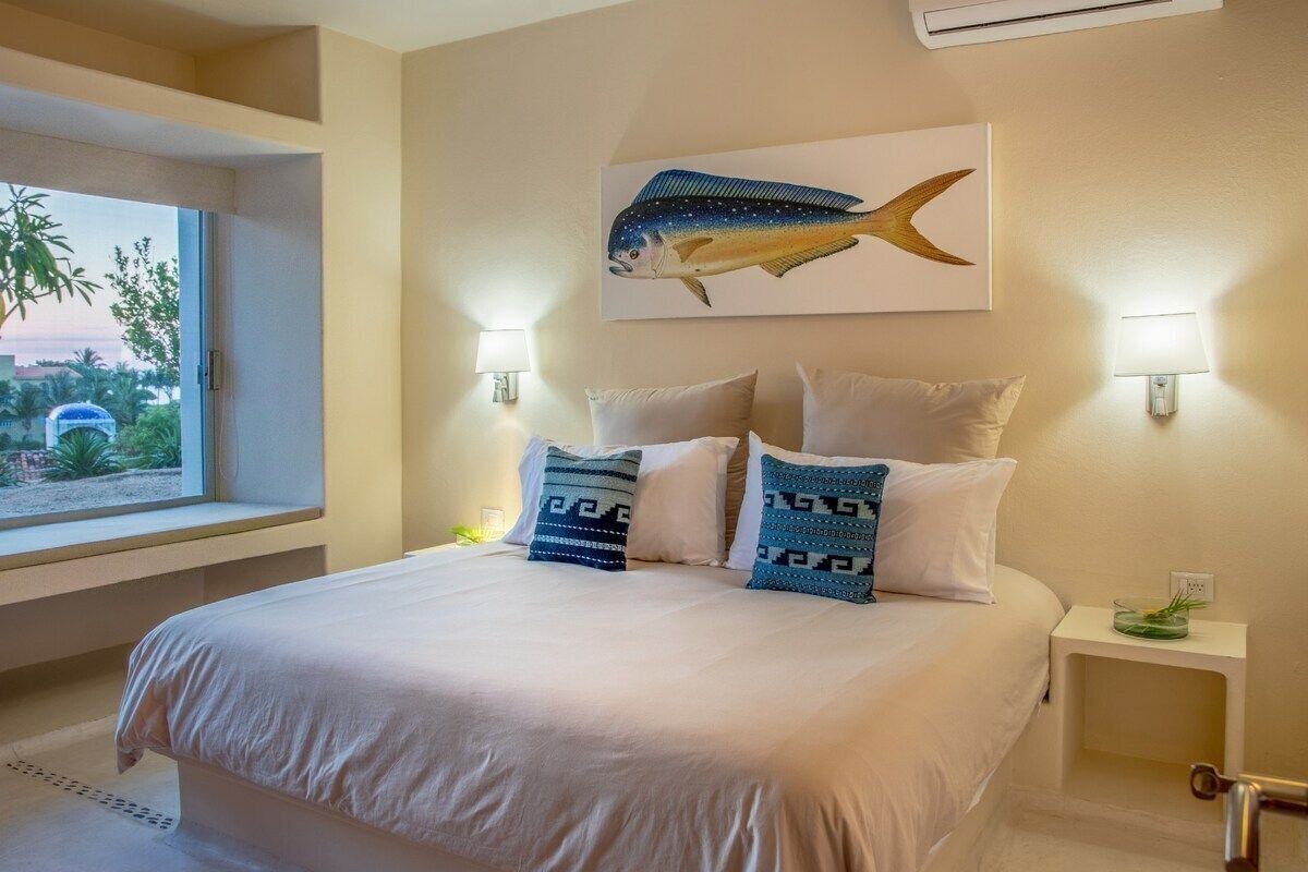 A hotel room with a large bed, two wall-mounted lamps, a painting of a fish, a window with a view of palm trees, and a small table with a plant.