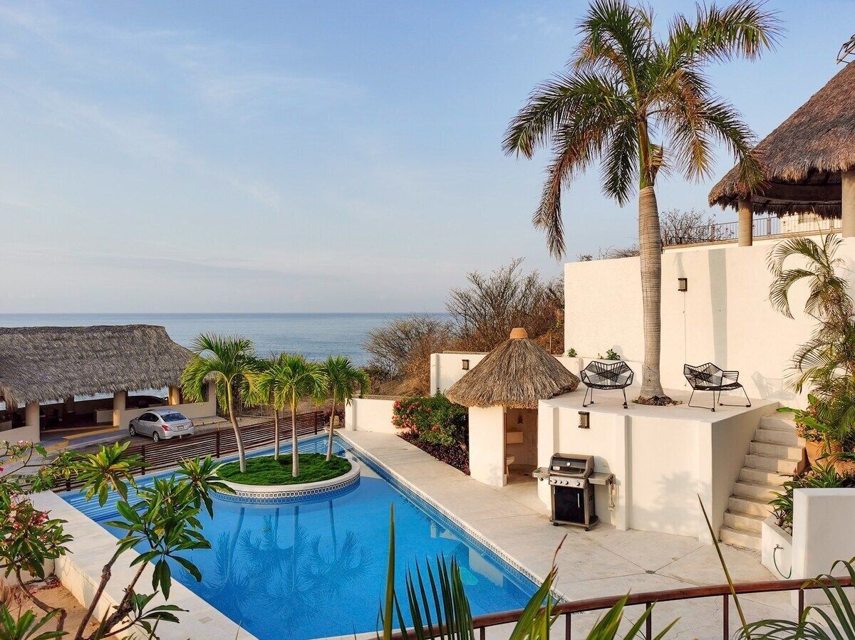 A pool area with a view of the sea, a barbecue setup, and a thatched-roof structure.