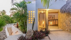 A modern building with a stone exterior, a well-maintained garden with palm trees, and a staircase leading to an interior with a stone wall and a hanging light fixture.