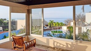 A balcony with a view of a pool and palm trees, wooden furniture, and a thatched umbrella.