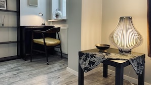 A room with a wooden floor, a black chair with a yellow cushion, a table with a black tablecloth and a lamp, a shelf with framed pictures, and a window with a plant. - Apartment next to charming Vistula Boulevards, Pekin (Kraków)