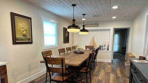 A dining area with a large wooden table and chairs, pendant lighting, and a view of the kitchen. - The CREW House at Bass Lake, IN (Bass Lake)