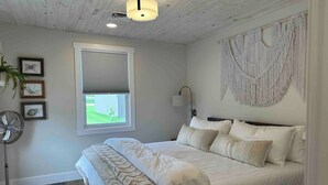 A bedroom with a bed, a window with blinds, a wall-mounted macrame piece, and a floor fan. - The CREW House at Bass Lake, IN (Bass Lake)