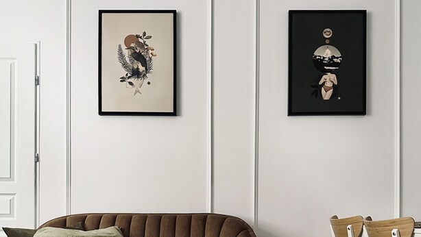 Un salon moderne avec un canapé marron, une table basse ronde et deux chaises blanches. Deux œuvres d’art encadrées ornent le mur.