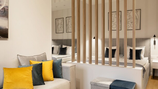 Une chambre d’hôtel moderne avec un grand lit, un canapé et une petite table. Les murs sont ornés de peintures abstraites et une cloison en bois sépare l’espace.