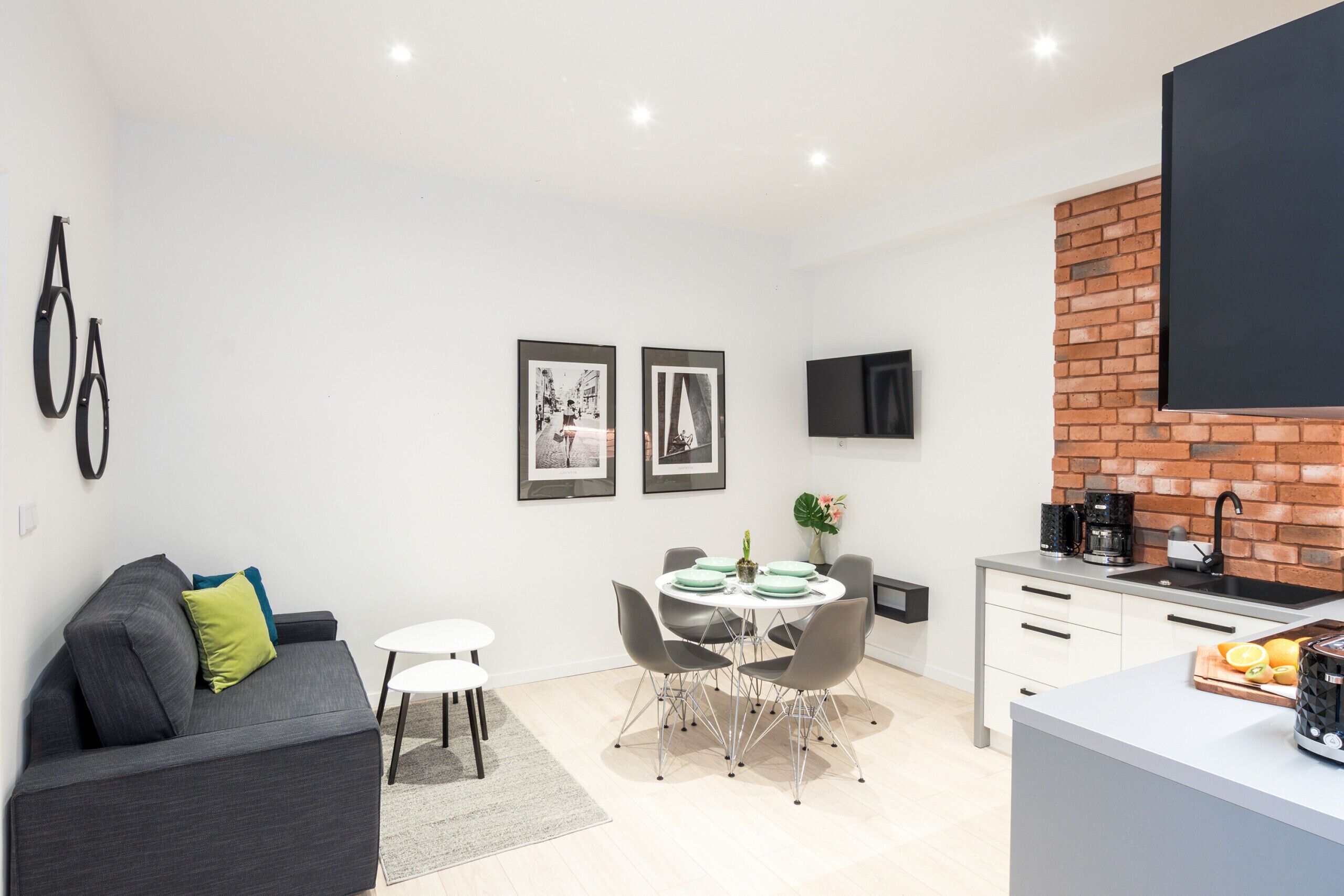 Un espace de vie moderne comprenant un canapé gris, une table à manger avec des chaises, un coin cuisine avec un mur de fond en briques et un téléviseur à écran plat fixé au mur.