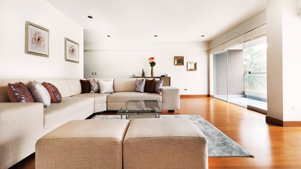 A modern living room with a sectional sofa, a glass coffee table, and a large window offering a view of the outdoors. - Beautiful 2,150 sq. ft. 3BR apartment in upscale Chacarilla (Lima)