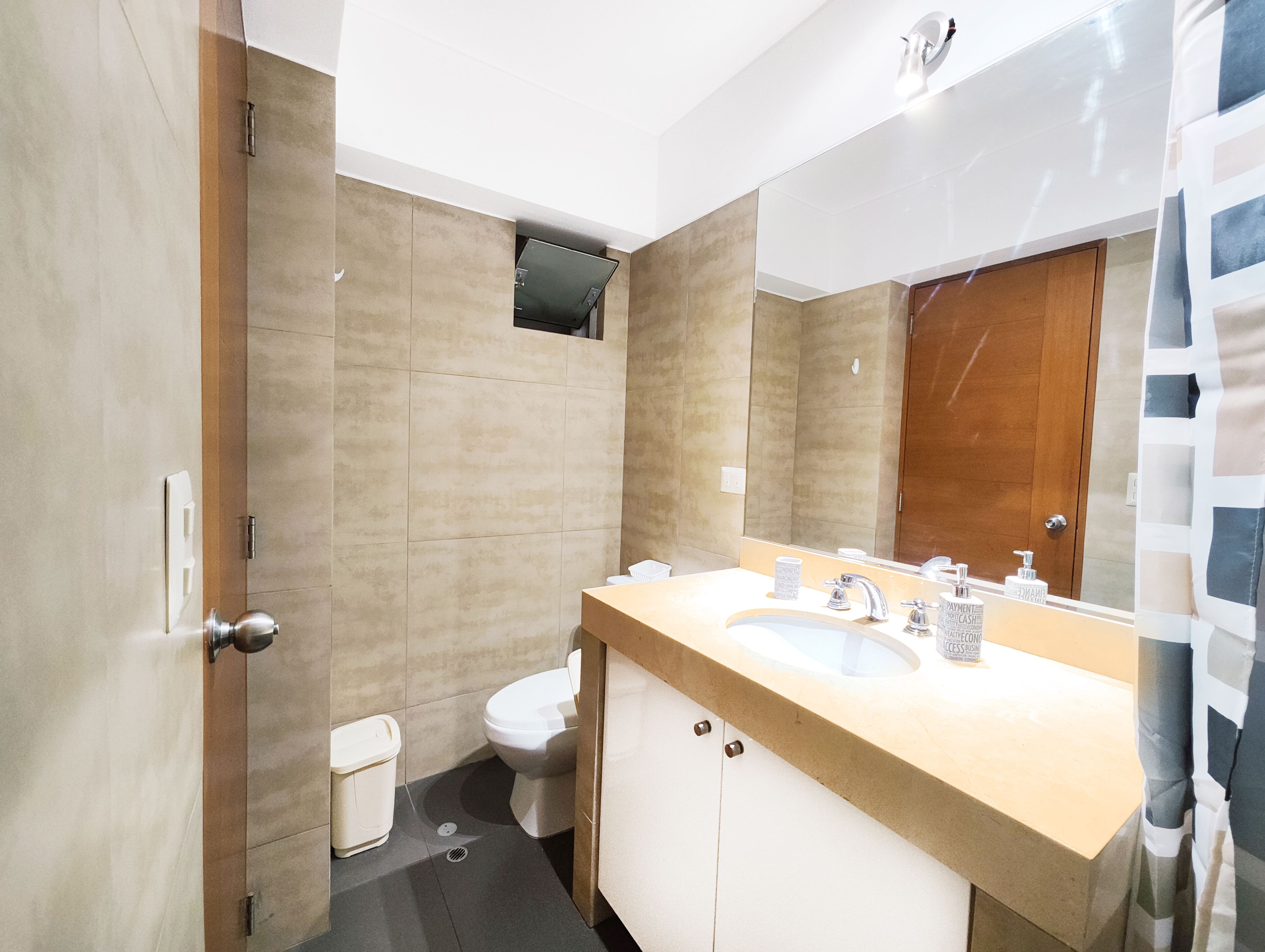 A modern bathroom with a beige tiled wall, a white toilet, a sink with a mirror, and a shower curtain.