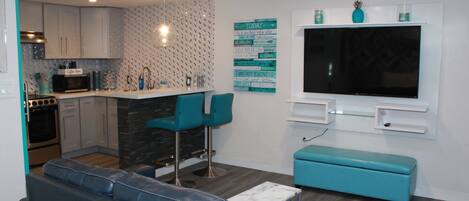 Cocina moderna con armarios blancos, microondas y cafetera. En la zona de estar, hay un banco color turquesa y un sofá azul, además de un televisor grande montado en la pared.