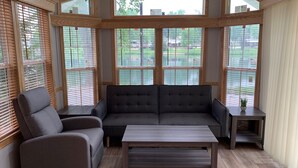 Ein modernes Wohnzimmer mit einem grauen Ecksofa, einem grauen Sessel und einem Holztisch. Große Fenster mit Holzrahmen und Jalousien sind sichtbar.