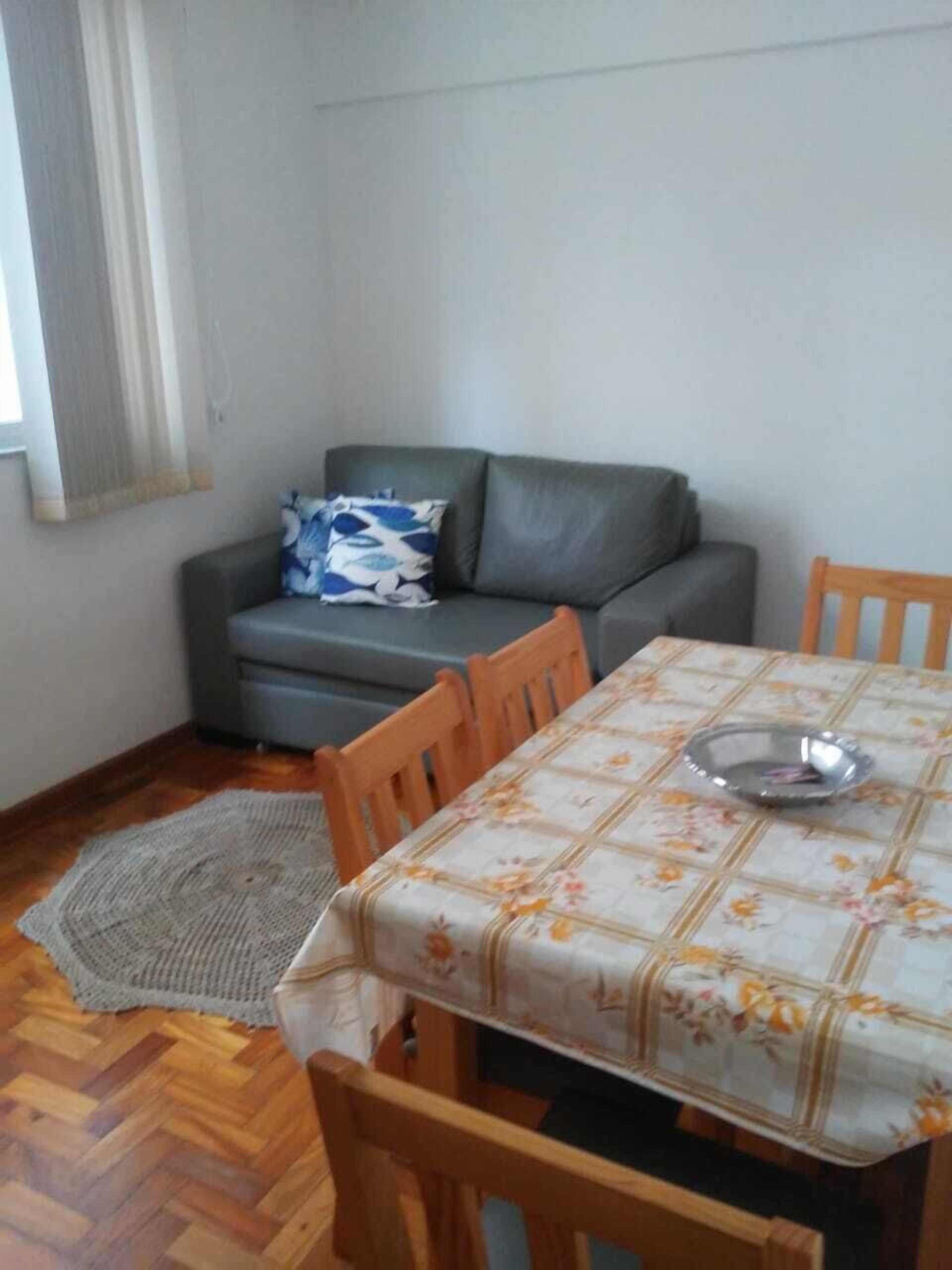 Ein Essbereich mit einem Tisch, der mit einer blumigen Tischdecke bedeckt ist, umgeben von Holzsesseln. Ein graues Sofa mit einem gemusterten Kissen lehnt an der Wand.