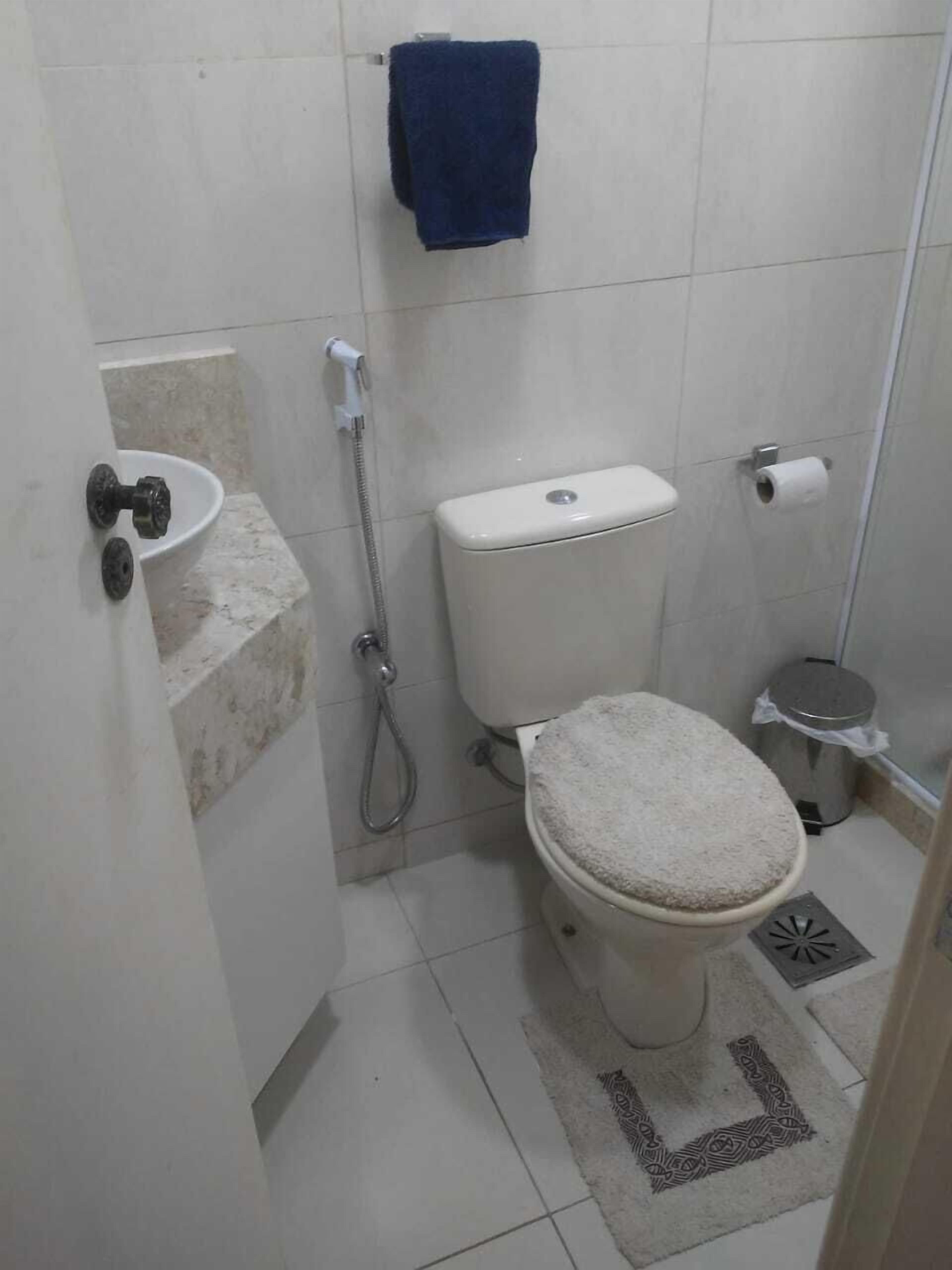 Casa de banho pequena e simples, com sanita, chuveiro, porta-toalhas e suporte para papel higiénico.