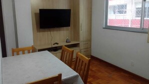 Un coin repas comprenant une table, des chaises, un téléviseur fixé à un meuble en bois et une fenêtre donnant sur un immeuble.