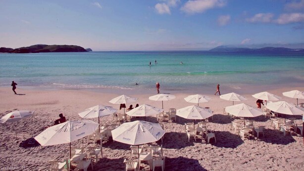 Une plage avec des parasols et des chaises de couleur blanche, une eau d’un bleu limpide et des gens qui se baignent.