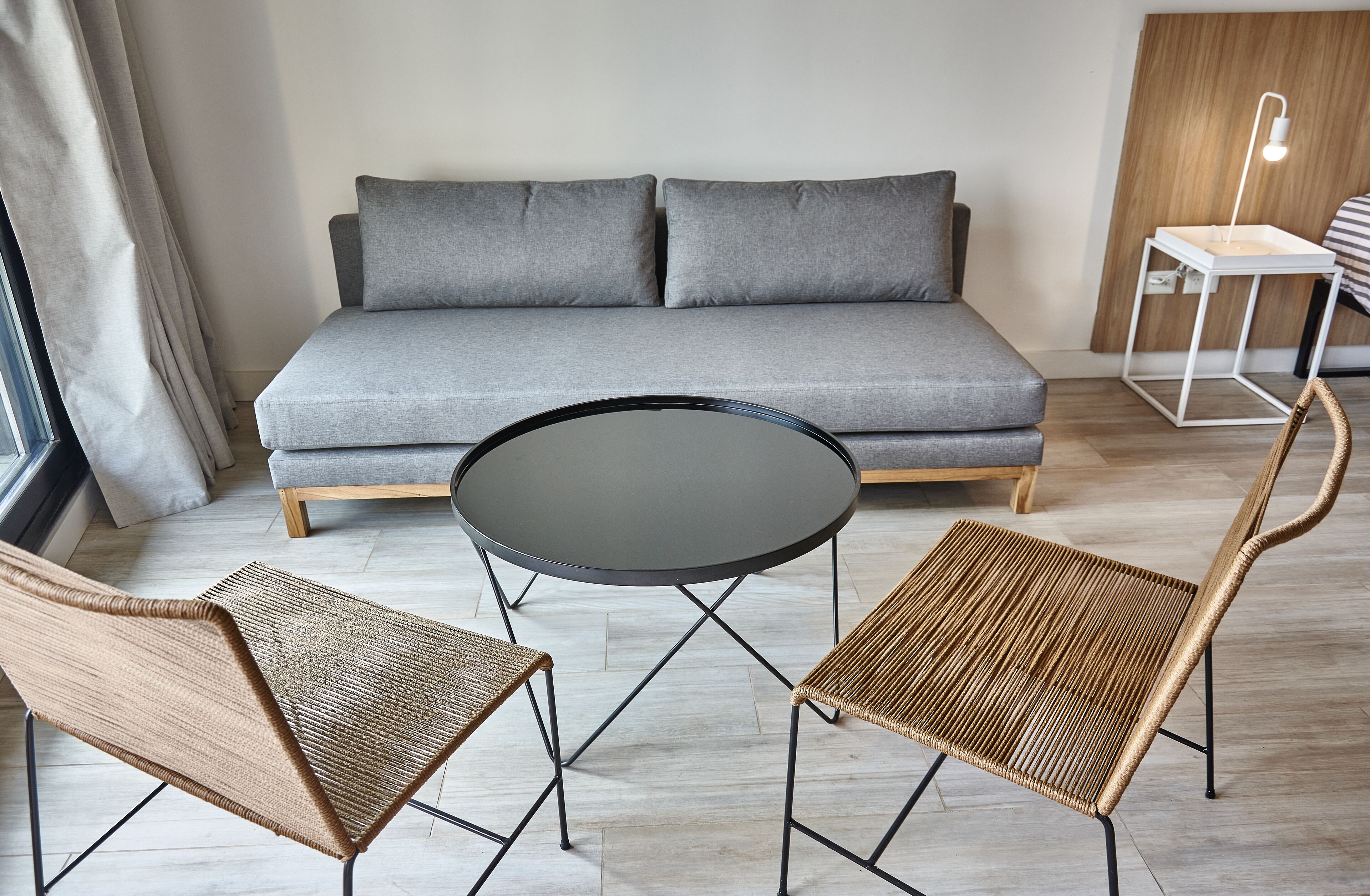 A modern living room with a grey sofa, two wicker chairs, a round table, and a bedside lamp.