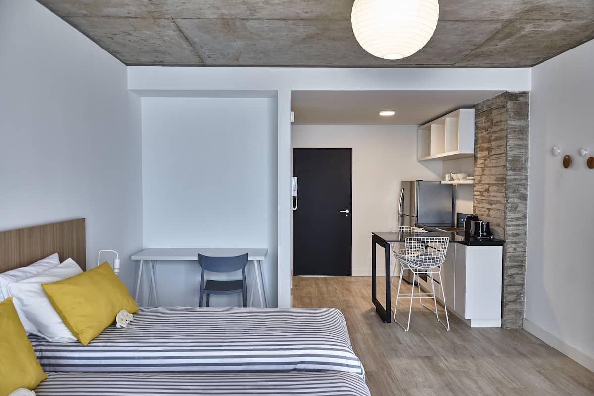 A modern hotel room with a bed, a desk, a chair, a kitchenette, and a door.