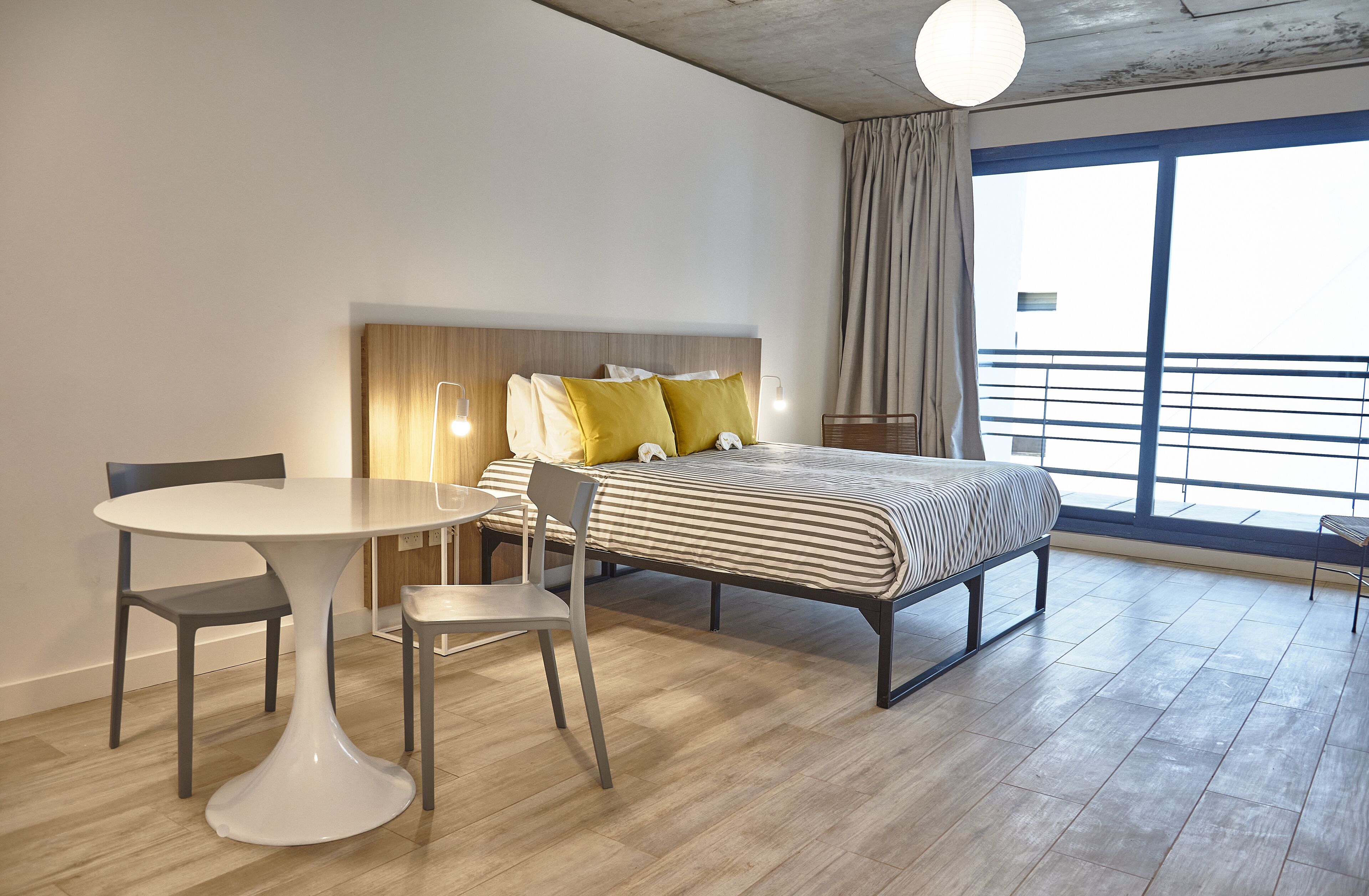A modern hotel room with a bed, a round dining table, two chairs, and a large window with a view.
